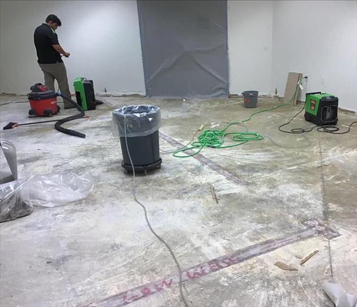 flooring removed exposing concrete floors and multiple pieces of drying equipment set up with a man checking their progress